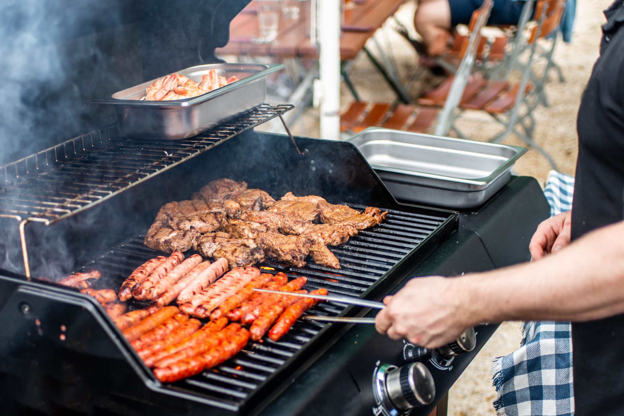 Person cooking brats and meat on an outdoor gas grill at a backyard cookout while preparing for summer grilling season